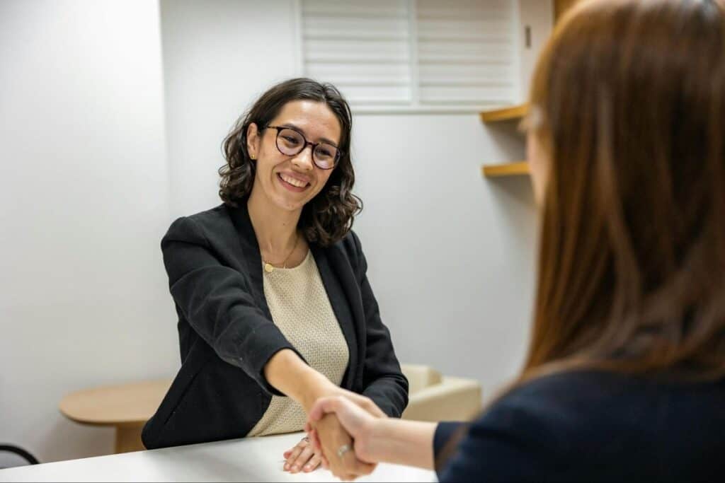 Two women shaking hands across a desk during a job interview, both smiling.