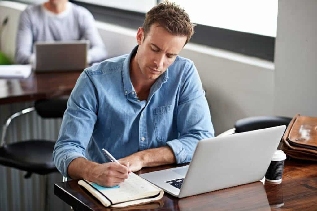Man writing on book and laptop in modern office for information with project