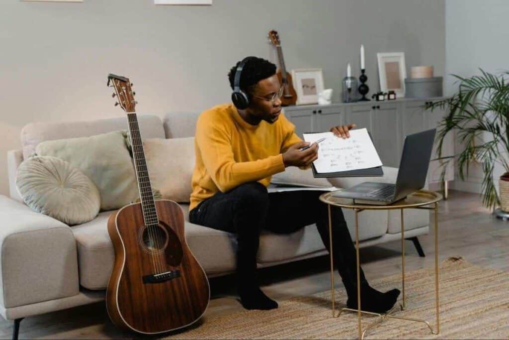 Imagen de archivo de un hombre dando clases particulares en línea con una guitarra, usando auriculares y un ordenador portátil para dar clases virtuales de música.