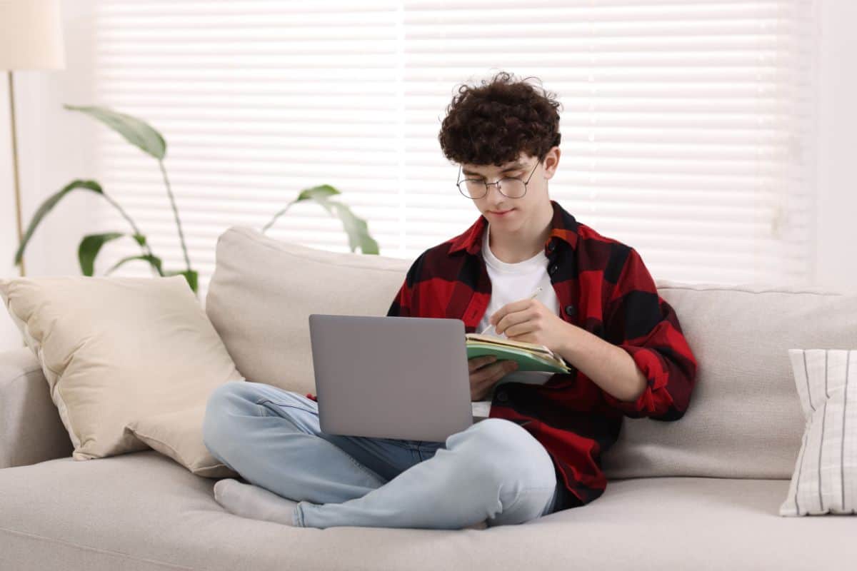 Teenager taking notes while working with laptop on sofa at home remote job