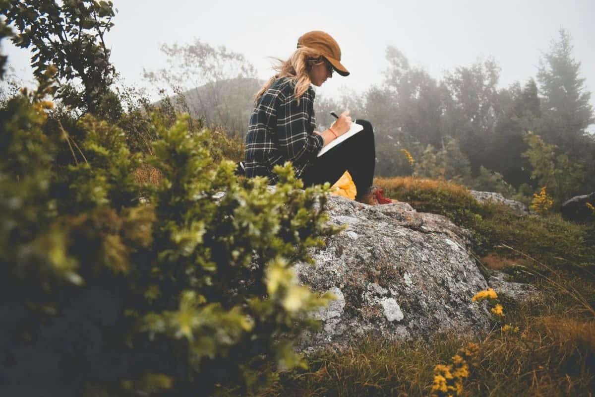 Stock image of a woman writing while seated on a large rock surrounded by trees in a natural outdoor setting