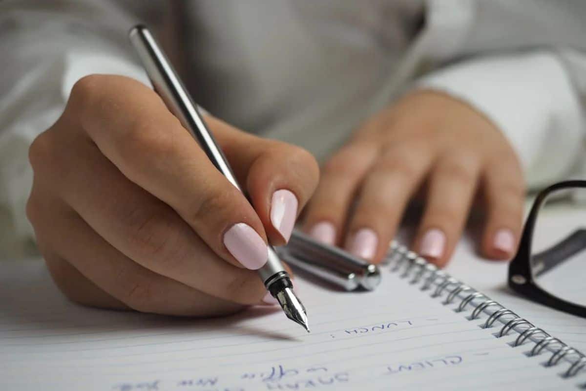 Woman in White Long Sleeved Shirt Holding a Pen Writing on a Paper