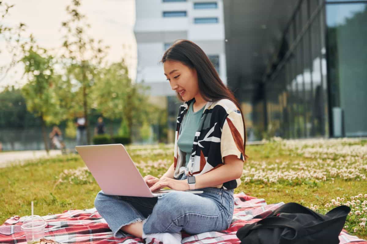 Sits with laptop young asian woman is outdoors at daytime