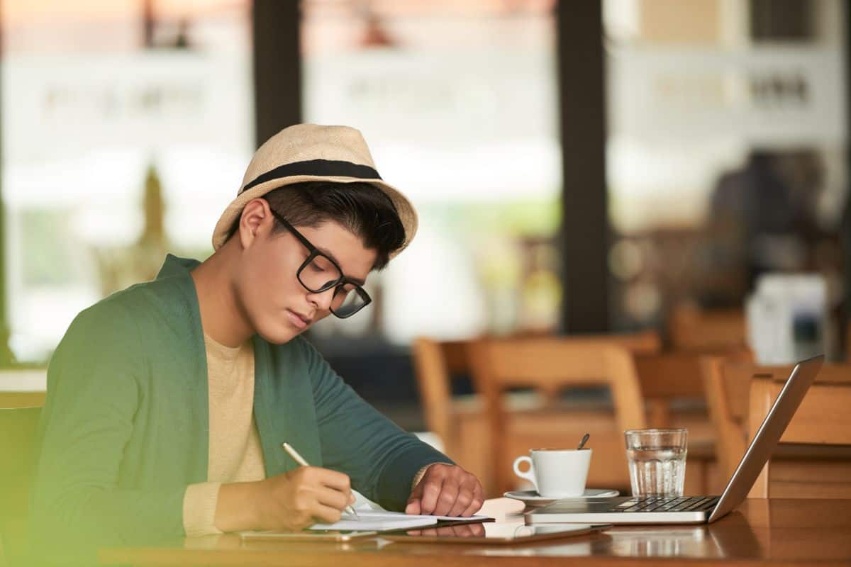 Giovane uomo asiatico alla moda seduto in un caffè con un computer portatile e scrivendo su un taccuino