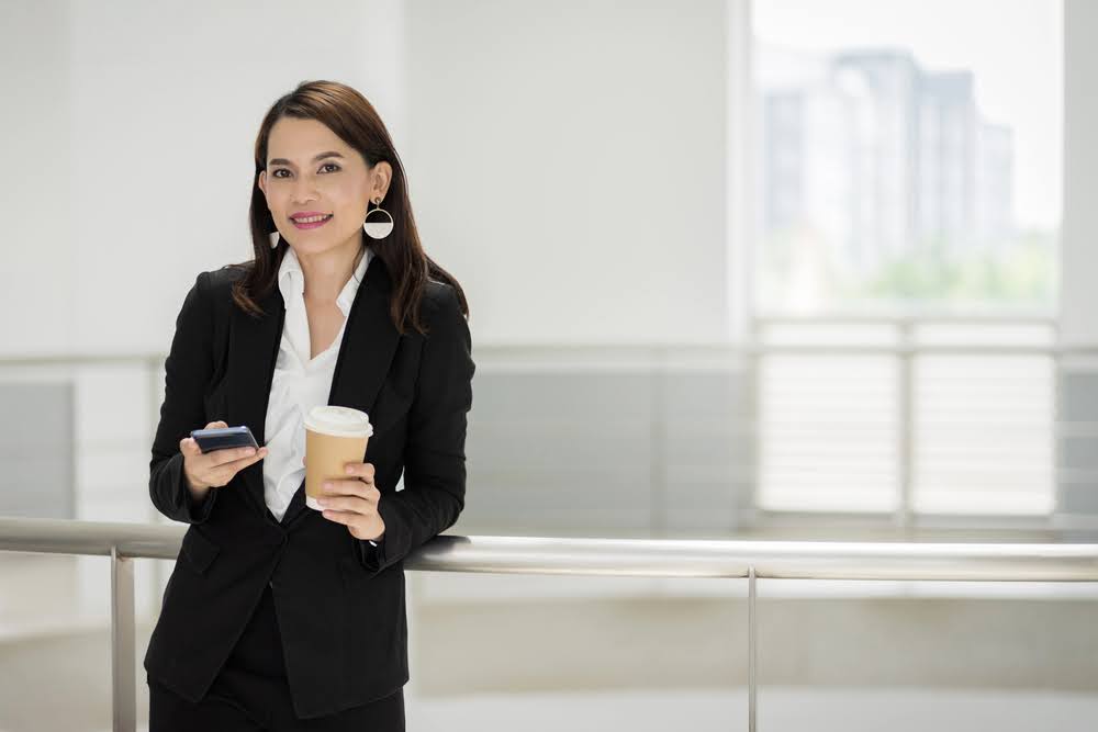 O que é um traje profissional de negócios (com exemplos) 10 Retrato de uma mulher de negócios confiante em um terno de negócios segurando uma xícara de café enquanto usa o celular durante o intervalo em um prédio comercial. Foto de estoque de negócios.