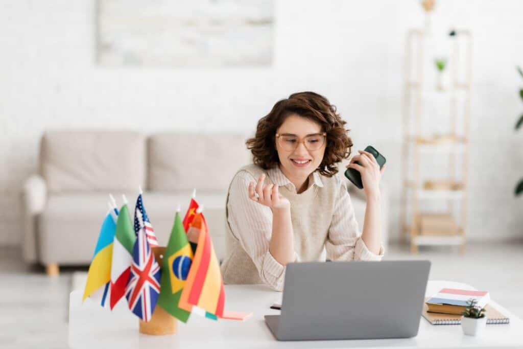 Cheerful language teacher in glasses holding smartphone near laptop 