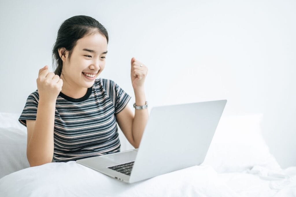 A woman wearing a striped shirt on the bed and playing laptop happily