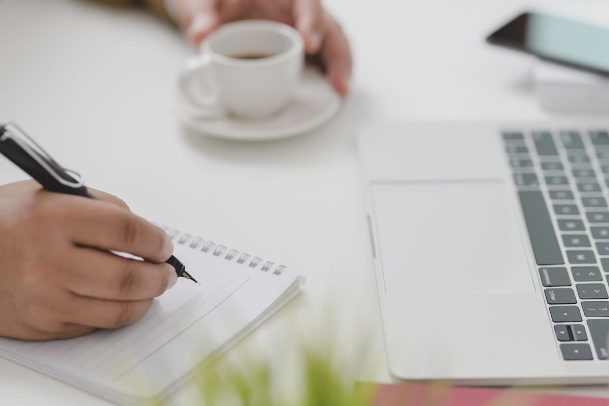 Person taking notes in a notebook beside a laptop