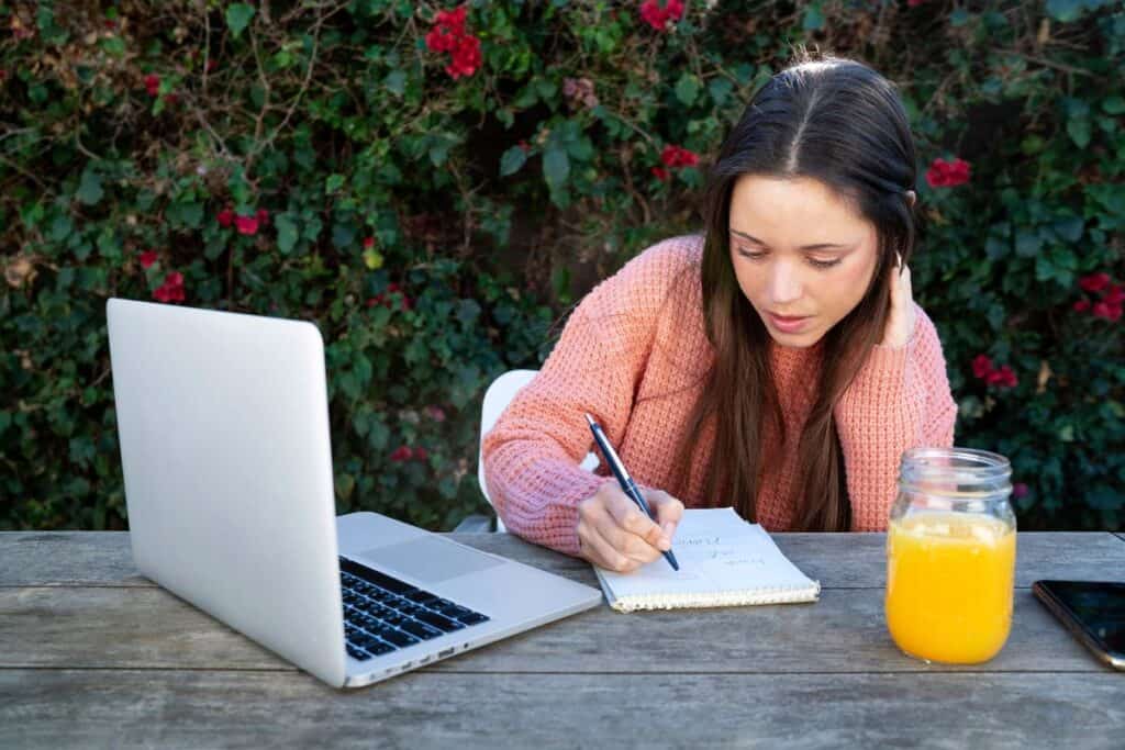 Como iniciar um blog: Tudo o que você precisa saber 8 Mulher jovem fazendo anotações ao ar livre enquanto usa um laptop