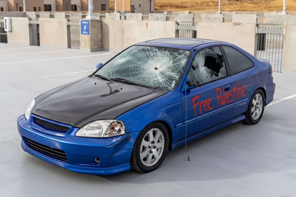 An AI-altered photo of the blue car, showing a cracked windshield, and spraypaint damage on the side of the car