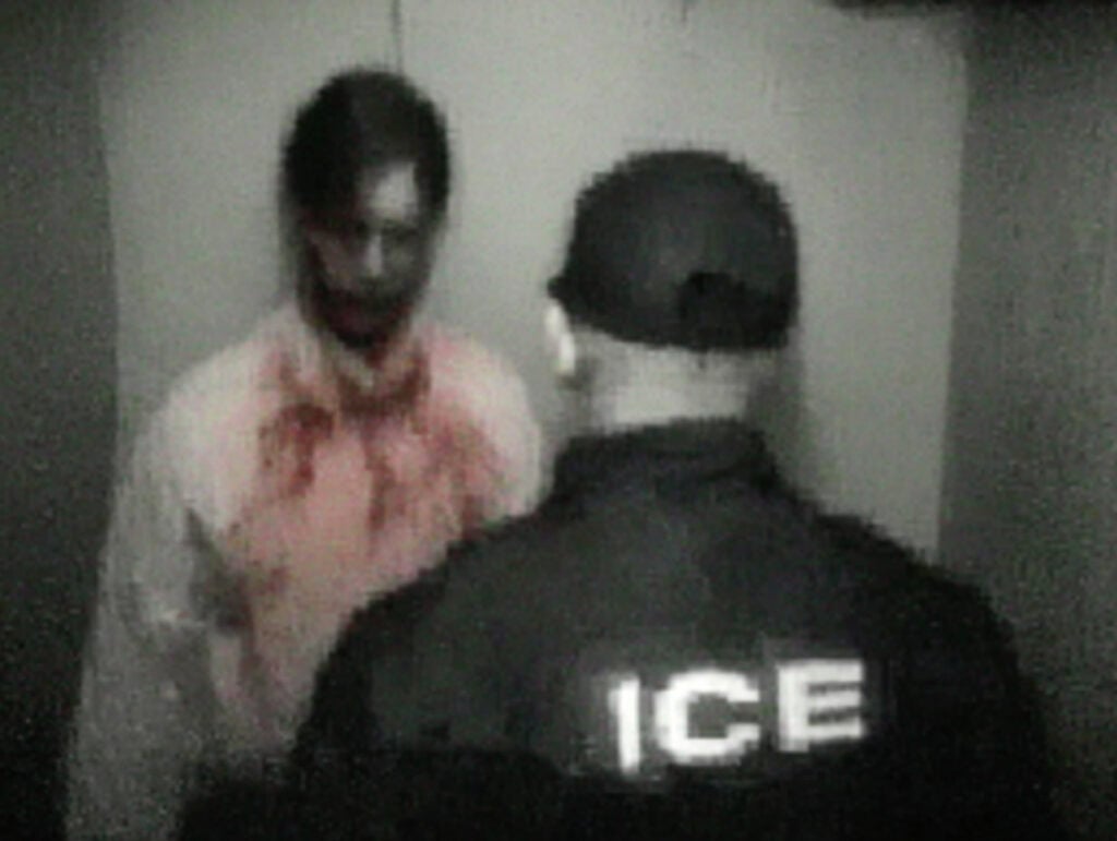 A blurry photo showing a man with a red-stained shirt, standing in front of someone.